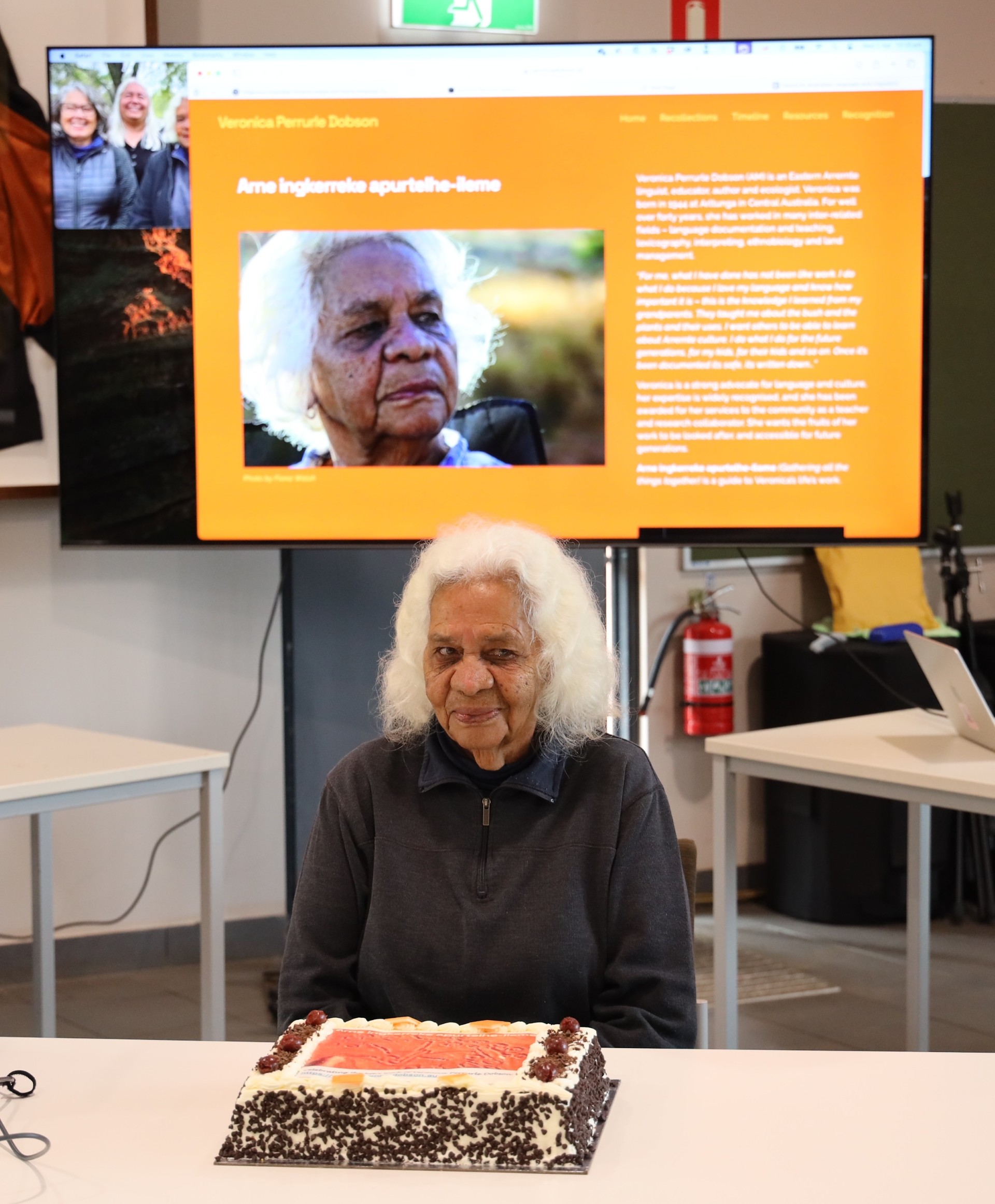 Veronica Perrurle Dobson celebrates the launch of Arne ingkerreke apurtelhe-ileme. A photograph of Veronica Perrurle Dobson with a celebratory cake. In the background is a large screen projecting the Arne ingkerreke apurtelhe-ileme website homepage.